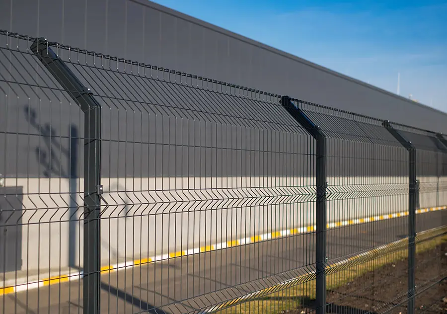 A wire fence runs alongside a paved road with a large industrial-looking gray building